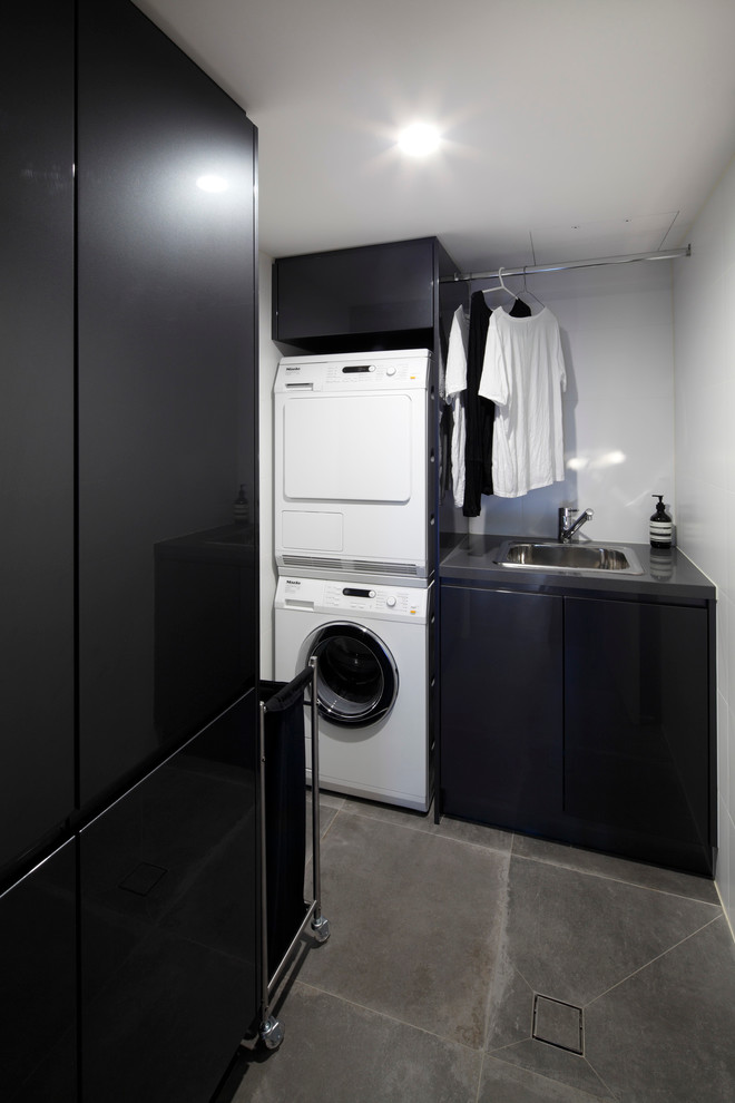 Inspiration for a mid-sized contemporary l-shaped dedicated laundry room in Sydney with recessed-panel cabinets, ceramic floors, a single-bowl sink, soapstone benchtops, white walls, a stacked washer and dryer and black cabinets.