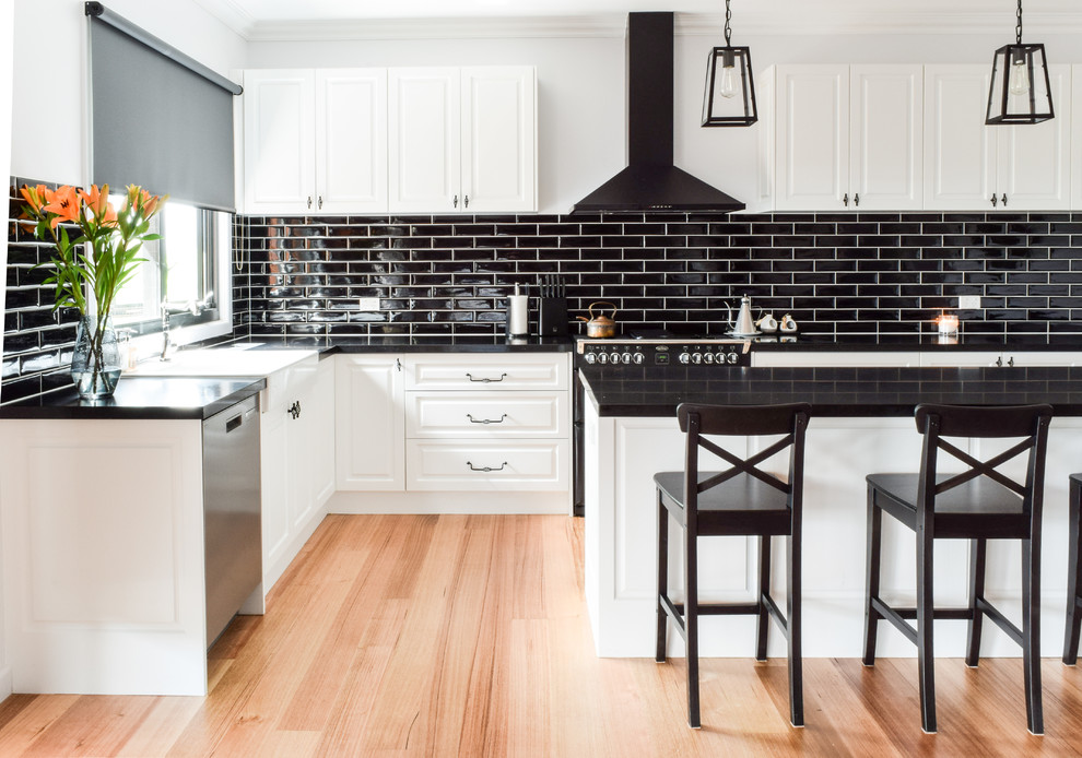 Photo of a large transitional l-shaped open plan kitchen in Melbourne with a farmhouse sink, quartz benchtops, black splashback, subway tile splashback, black appliances, with island, brown floor, raised-panel cabinets and medium hardwood floors.