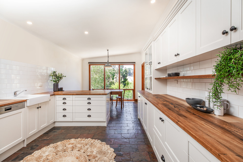 This is an example of a mid-sized country l-shaped eat-in kitchen in Adelaide with a farmhouse sink, shaker cabinets, white cabinets, wood benchtops, white splashback, subway tile splashback, black appliances, slate floors, a peninsula, grey floor and yellow benchtop.