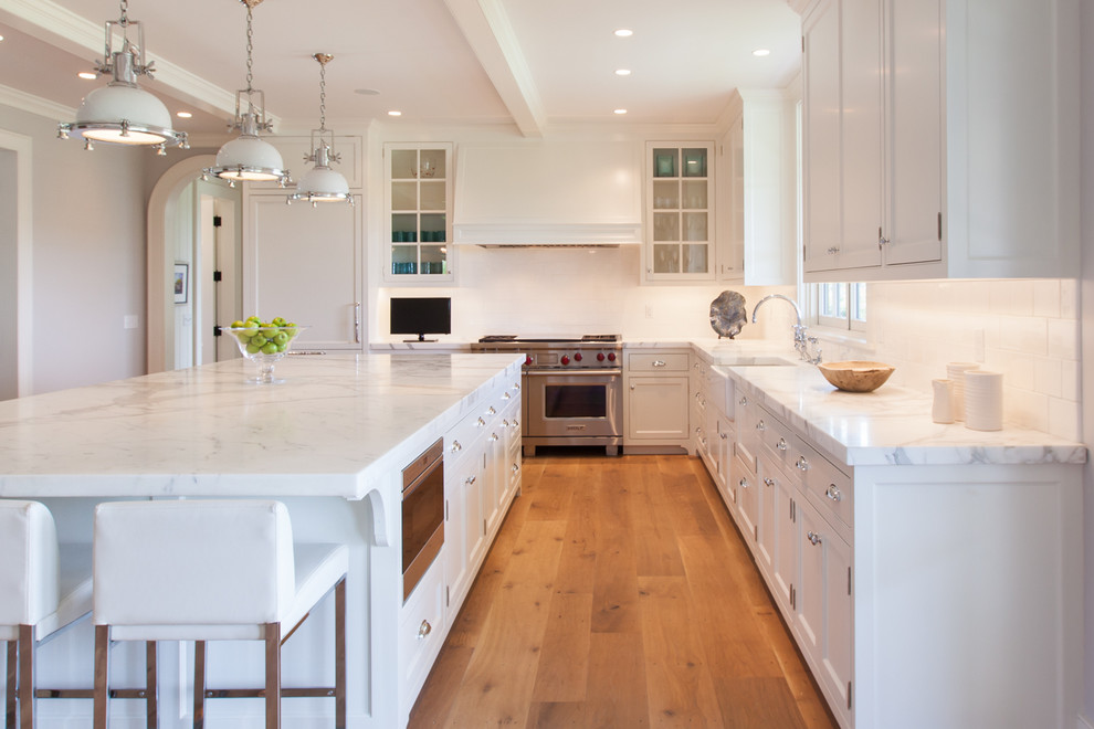 Inspiration for a large beach style l-shaped open plan kitchen in Boston with a farmhouse sink, glass-front cabinets, white cabinets, marble benchtops, white splashback, ceramic splashback, stainless steel appliances, medium hardwood floors and with island.