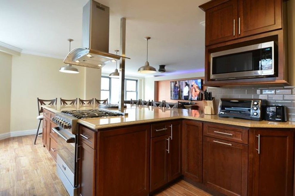 Photo of a large traditional galley eat-in kitchen in New York with a farmhouse sink, shaker cabinets, brown cabinets, granite benchtops, beige splashback, glass tile splashback, stainless steel appliances, light hardwood floors and a peninsula.