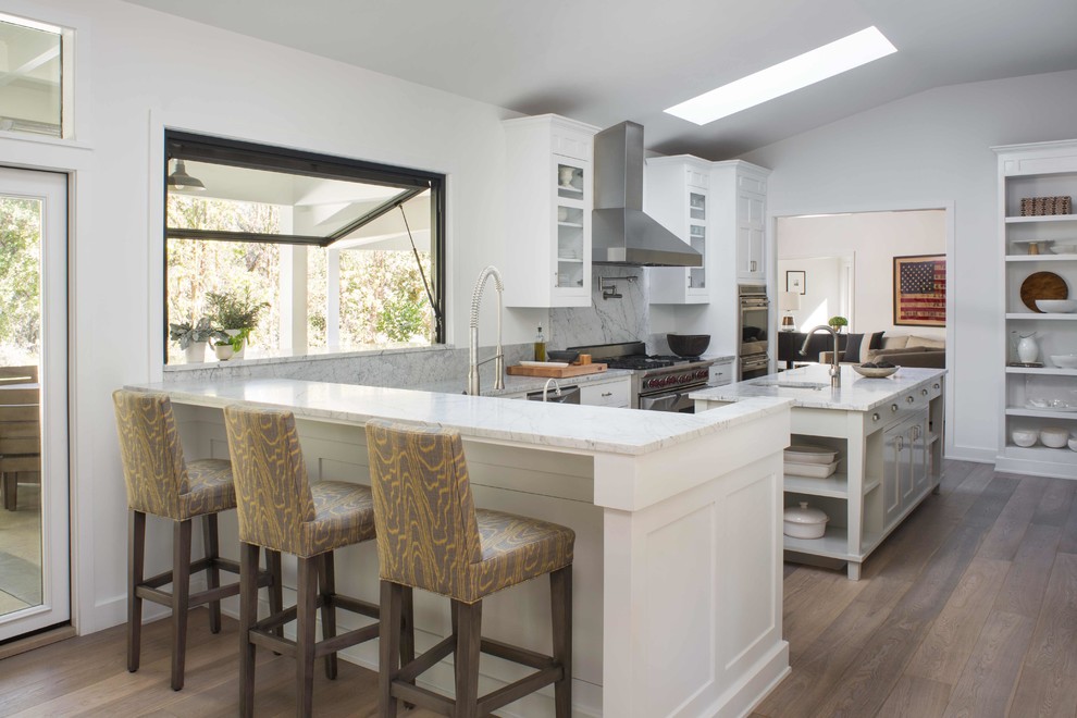Large country l-shaped separate kitchen in Los Angeles with a farmhouse sink, shaker cabinets, white cabinets, marble benchtops, white splashback, marble splashback, stainless steel appliances, light hardwood floors, with island and white benchtop.