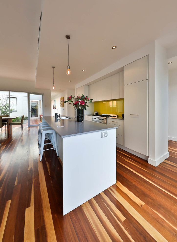 Photo of a mid-sized contemporary galley open plan kitchen in Adelaide with a double-bowl sink, white cabinets, quartz benchtops, yellow splashback, glass tile splashback, stainless steel appliances, medium hardwood floors, with island and flat-panel cabinets.