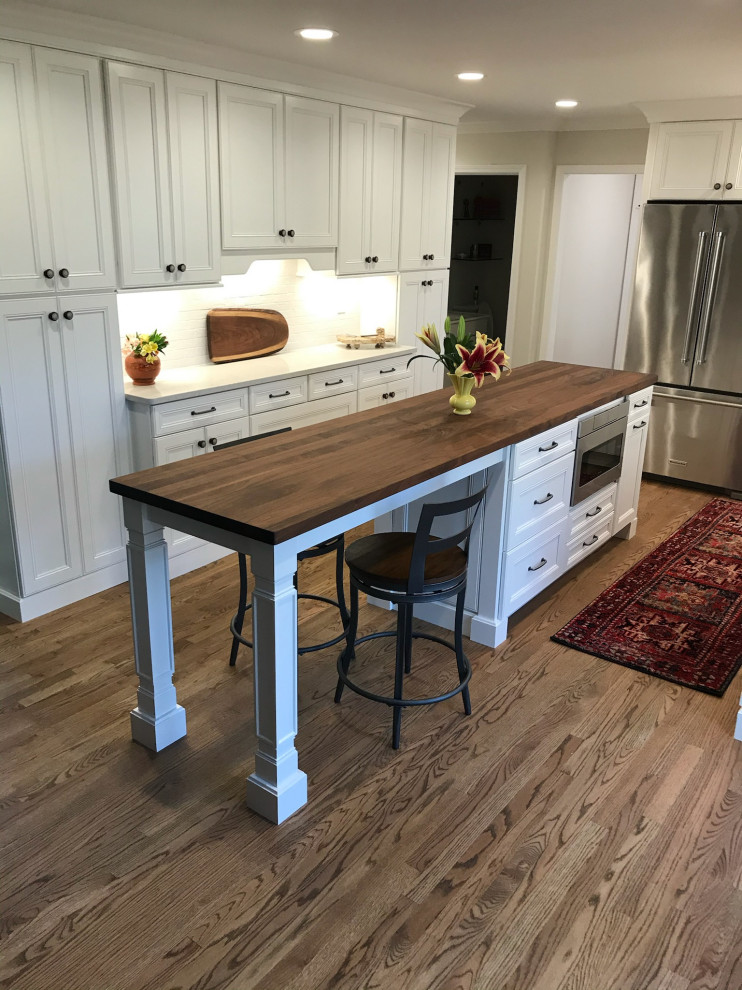 Example of a large transitional galley medium tone wood floor and brown floor eat-in kitchen design in Other with recessed-panel cabinets, white cabinets, quartz countertops, white backsplash, brick backsplash, stainless steel appliances, an island and white countertops