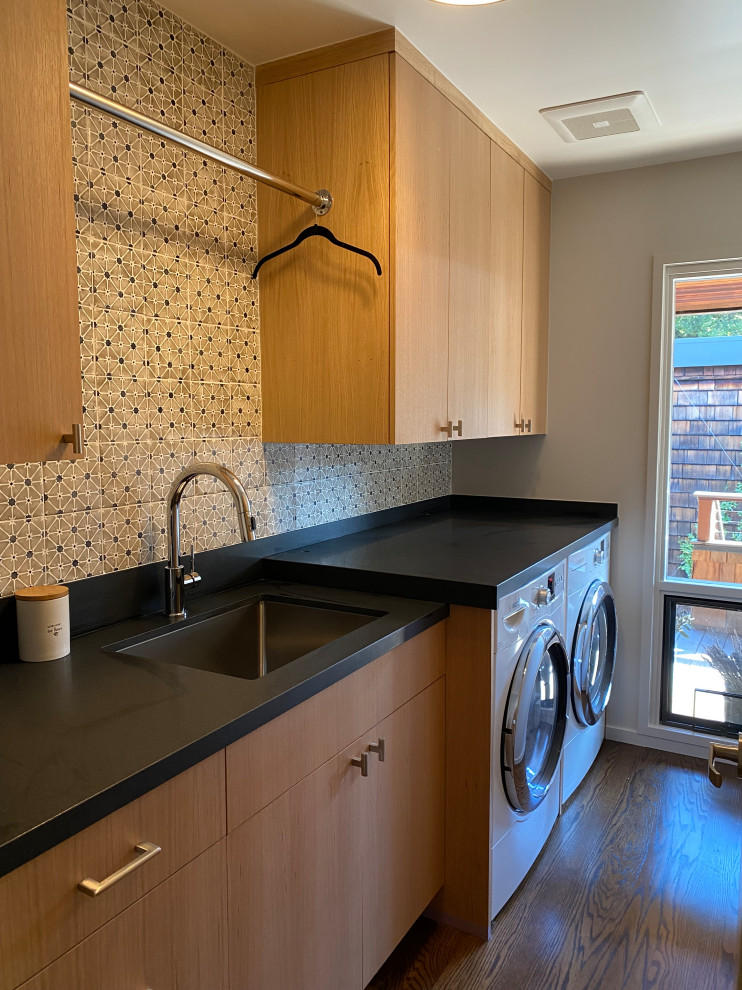 This is an example of a small modern single-wall dedicated laundry room in San Francisco with a single-bowl sink, flat-panel cabinets, light wood cabinets, soapstone benchtops, multi-coloured splashback, porcelain splashback, beige walls, medium hardwood floors, an integrated washer and dryer, brown floor and black benchtop.