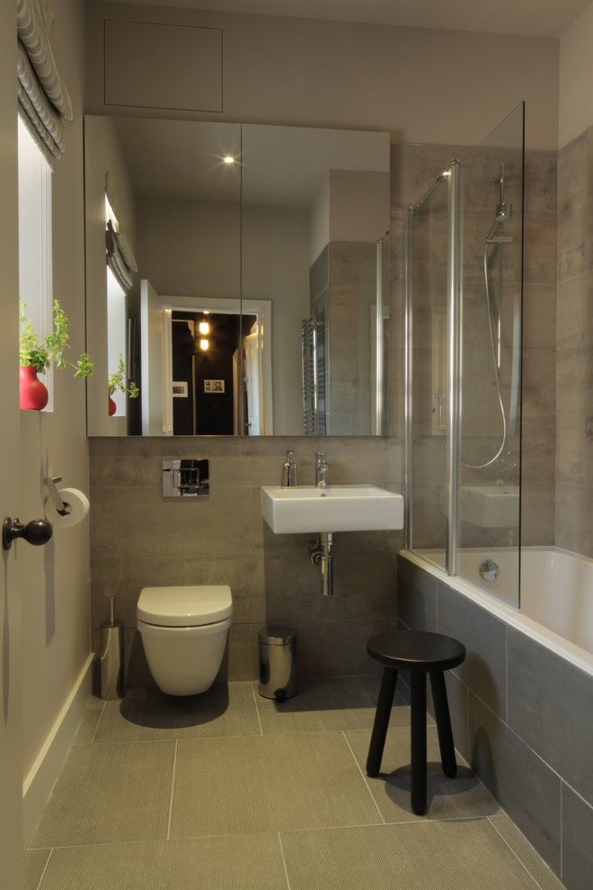 This is an example of a mid-sized contemporary kids bathroom in London with a drop-in tub, a shower/bathtub combo, a wall-mount toilet, gray tile, ceramic tile, grey walls, ceramic floors and a wall-mount sink.