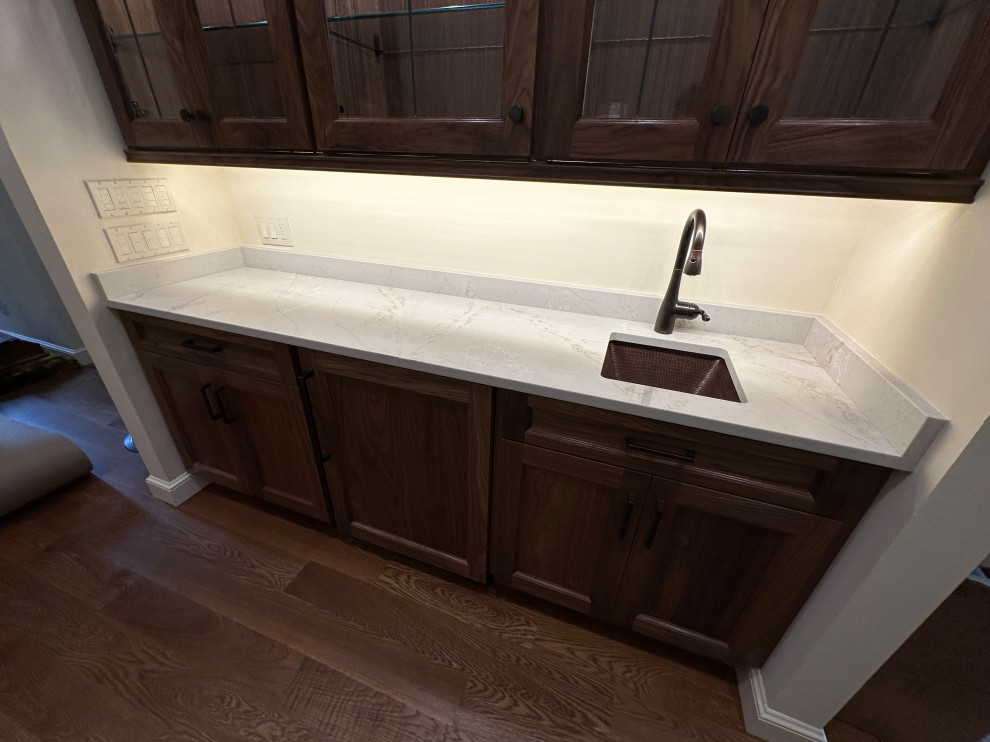 This is an example of a small traditional single-wall wet bar in Cleveland with an undermount sink, glass-front cabinets, dark wood cabinets, quartzite benchtops, white splashback, medium hardwood floors, brown floor and white benchtop.