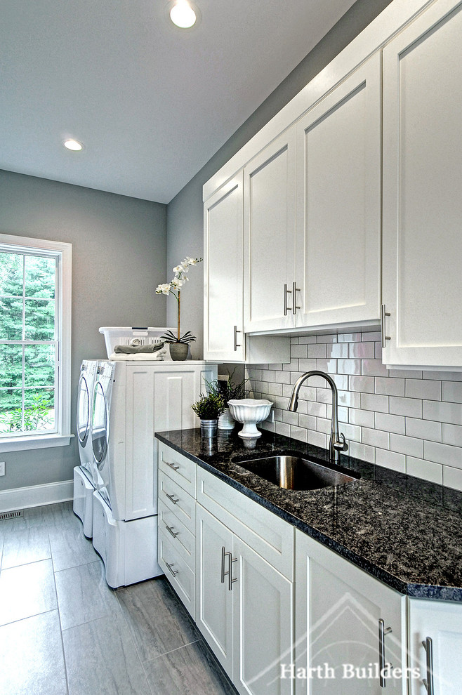 Inspiration for a large transitional galley utility room in Philadelphia with an undermount sink, shaker cabinets, white cabinets, granite benchtops, grey walls, porcelain floors, a side-by-side washer and dryer, grey floor and black benchtop.