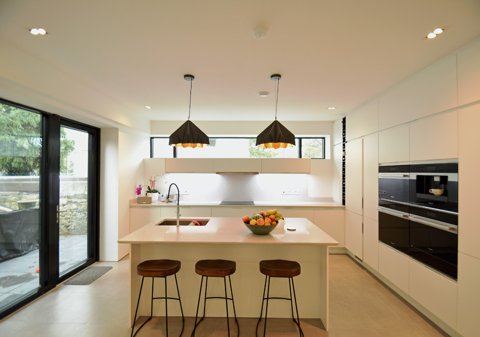 Photo of a mid-sized contemporary l-shaped open plan kitchen in Cork with an integrated sink, flat-panel cabinets, white cabinets, quartz benchtops, white splashback, ceramic splashback, black appliances, cement tiles, with island, beige floor and white benchtop.