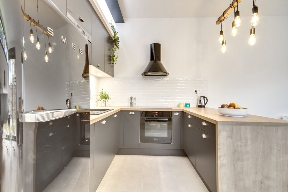 This is an example of a mid-sized industrial u-shaped open plan kitchen in Paris with laminate benchtops, white splashback, subway tile splashback, a single-bowl sink, flat-panel cabinets, grey cabinets, black appliances, ceramic floors, grey floor and brown benchtop.