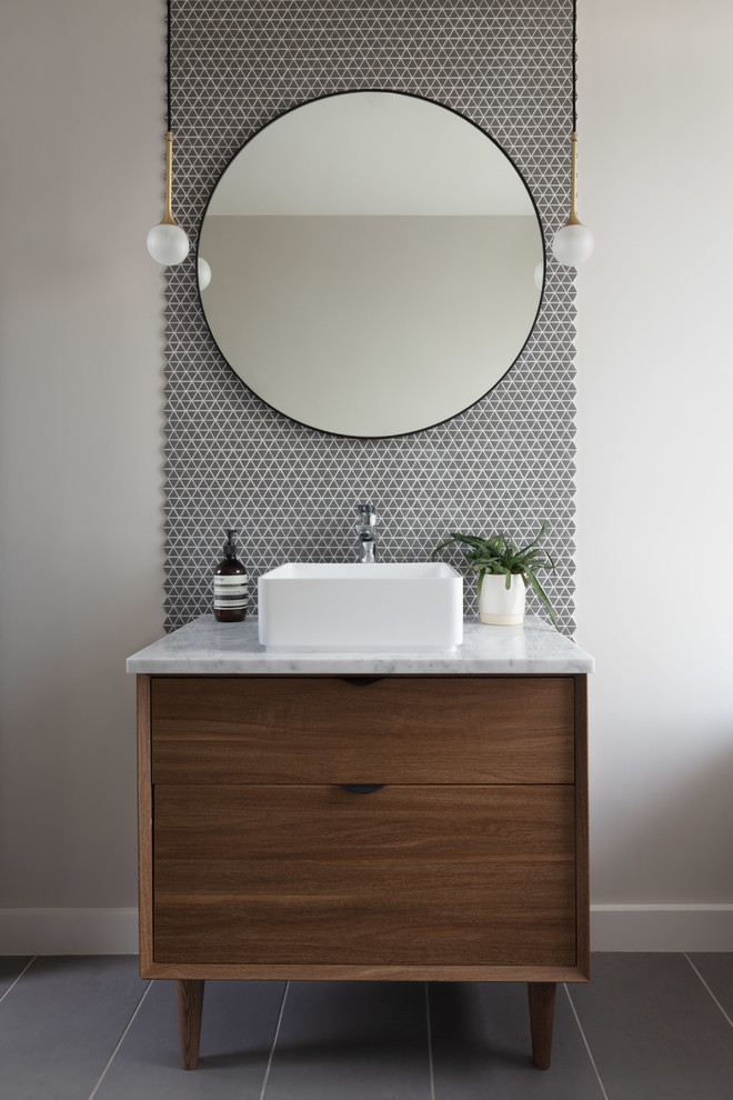 This is an example of a mid-sized contemporary kids bathroom in London with flat-panel cabinets, medium wood cabinets, a freestanding tub, gray tile, ceramic tile, grey walls, ceramic floors, a console sink, marble benchtops, grey floor and white benchtops.