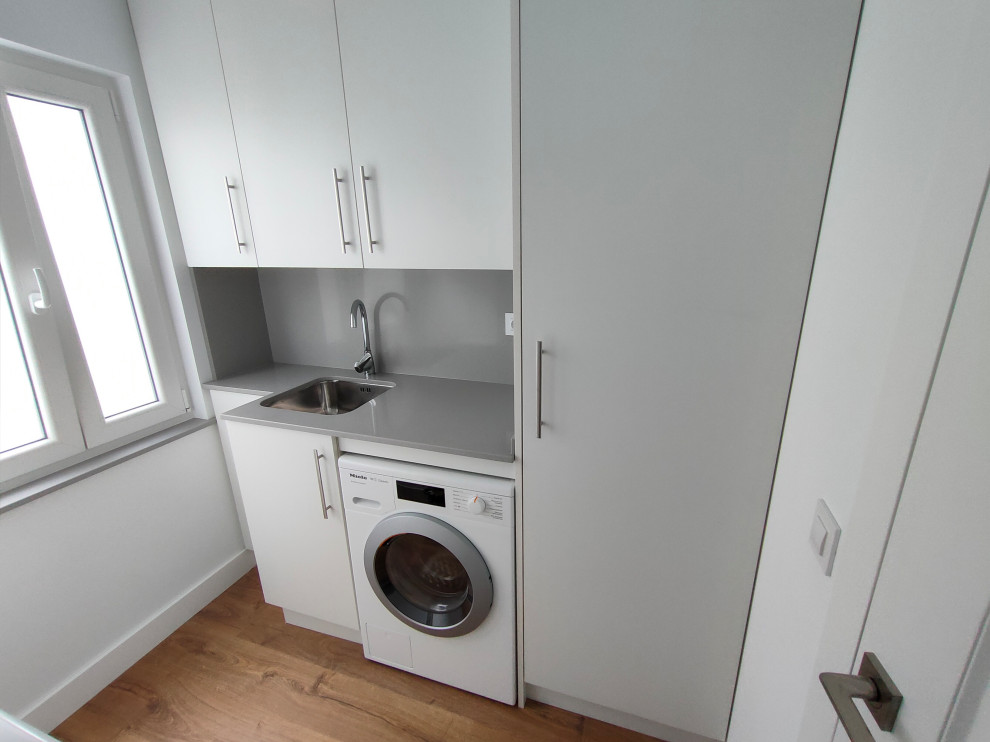 Design ideas for a mid-sized transitional single-wall dedicated laundry room in Other with an undermount sink, flat-panel cabinets, white cabinets, quartz benchtops, white walls, medium hardwood floors, an integrated washer and dryer, brown floor and grey benchtop.