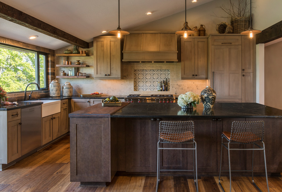 Photo of a mid-sized transitional l-shaped kitchen in Portland with a farmhouse sink, shaker cabinets, medium wood cabinets, soapstone benchtops, beige splashback, porcelain splashback, stainless steel appliances, medium hardwood floors, with island, brown floor and black benchtop.