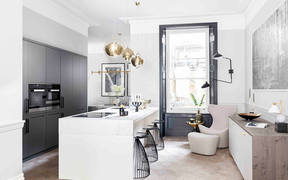 Contemporary kitchen in London with an undermount sink, flat-panel cabinets, grey cabinets, black appliances, light hardwood floors, with island and white benchtop.