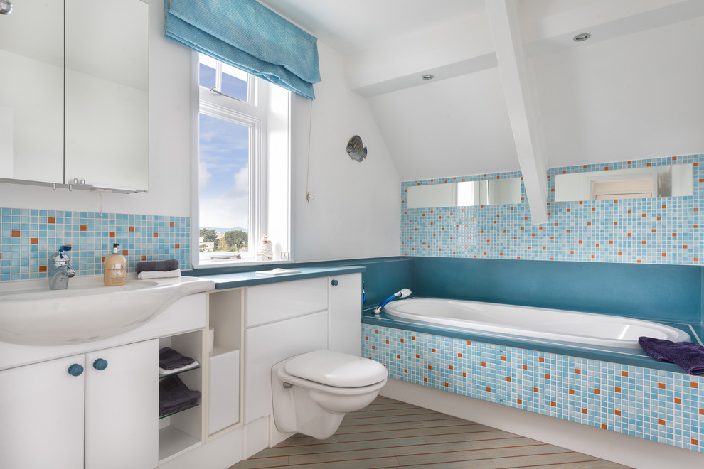 Photo of a mid-sized beach style 3/4 bathroom in Devon with a vessel sink, flat-panel cabinets, white cabinets, laminate benchtops, a drop-in tub, a wall-mount toilet, blue tile, ceramic tile, white walls and linoleum floors.