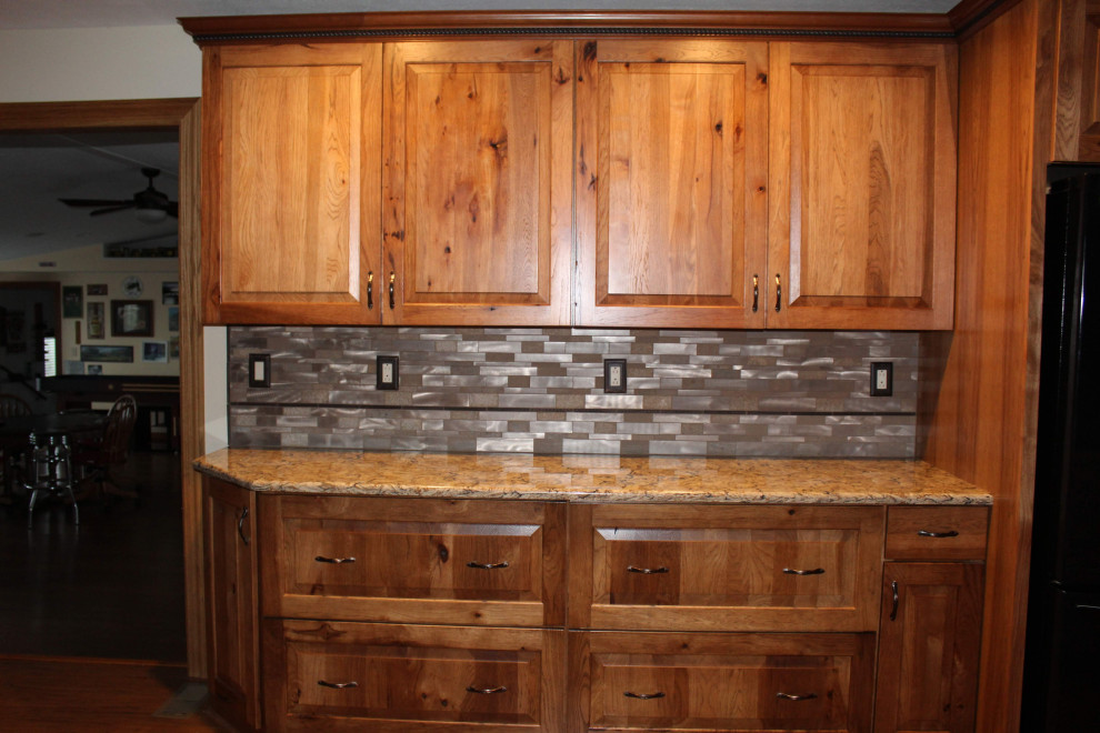 Large mountain style l-shaped medium tone wood floor and brown floor open concept kitchen photo with an undermount sink, raised-panel cabinets, brown cabinets, quartzite countertops, metallic backsplash, black appliances, an island and brown countertops