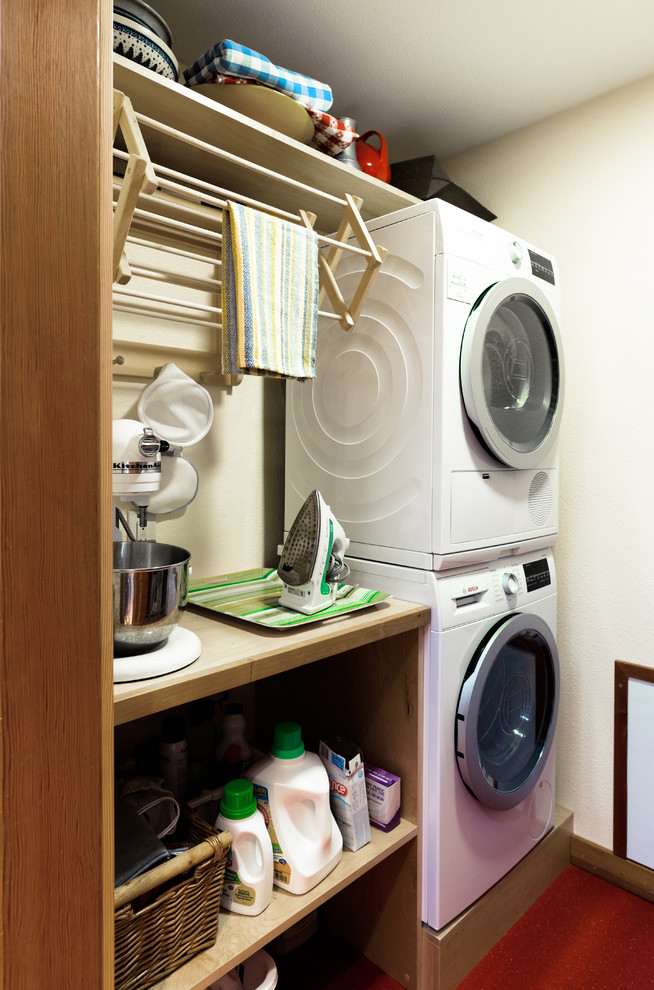This is an example of a small country galley utility room in Seattle with flat-panel cabinets, light wood cabinets, wood benchtops, linoleum floors, a stacked washer and dryer, red floor, white walls and beige benchtop.