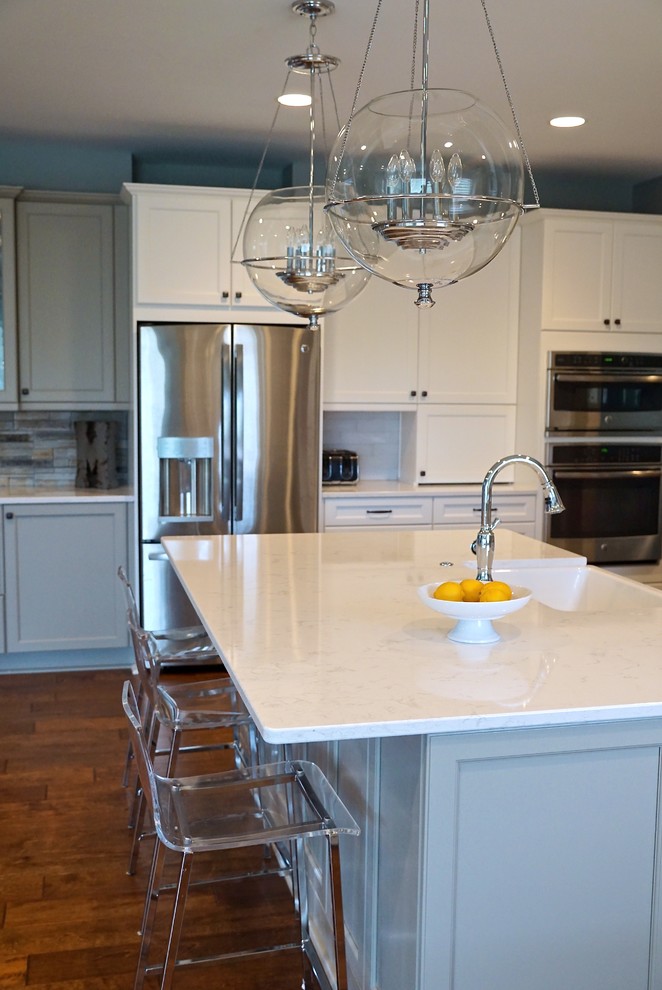 This is an example of a large transitional kitchen in Omaha with a farmhouse sink, shaker cabinets, white cabinets, quartz benchtops, white splashback, glass tile splashback, stainless steel appliances, medium hardwood floors and with island.