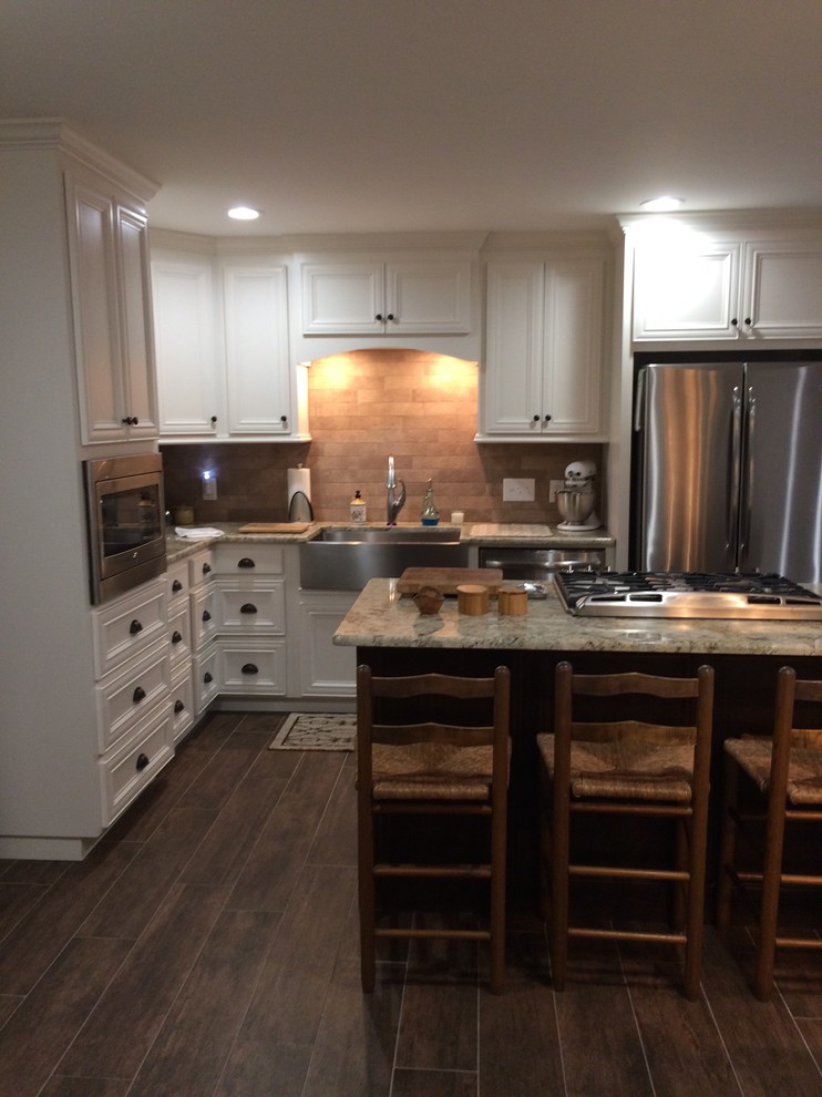 Example of a large transitional l-shaped brown floor eat-in kitchen design in Birmingham with a farmhouse sink, recessed-panel cabinets, white cabinets, beige backsplash, stainless steel appliances and an island
