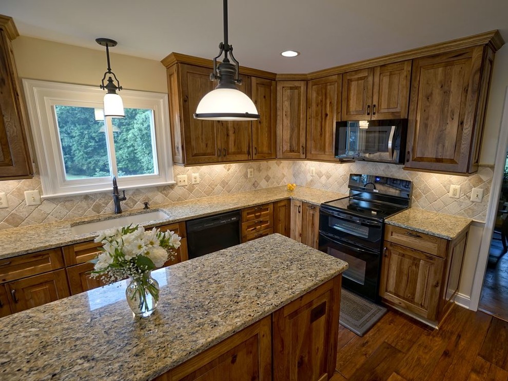 Photo of a mid-sized country l-shaped eat-in kitchen in DC Metro with an undermount sink, raised-panel cabinets, brown cabinets, granite benchtops, beige splashback, travertine splashback, black appliances, dark hardwood floors, with island, brown floor and beige benchtop.