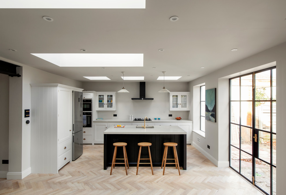 This is an example of a mid-sized transitional l-shaped open plan kitchen in London with shaker cabinets, concrete benchtops, light hardwood floors, with island, grey benchtop, white cabinets, stainless steel appliances and beige floor.