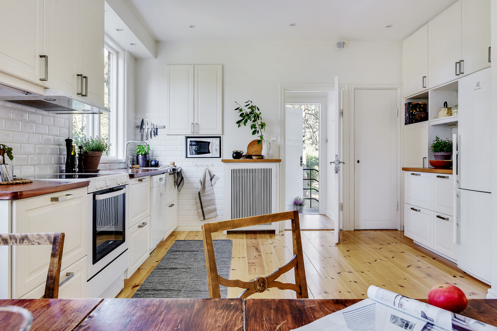 Inspiration for a scandinavian galley eat-in kitchen in Stockholm with an undermount sink, raised-panel cabinets, white cabinets, wood benchtops, white splashback, subway tile splashback, white appliances, light hardwood floors, brown floor and brown benchtop.