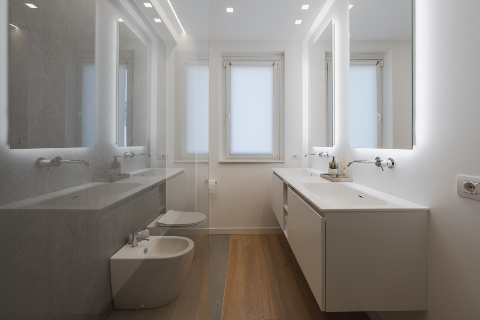 This is an example of a mid-sized contemporary 3/4 bathroom in Milan with flat-panel cabinets, white cabinets, an open shower, a two-piece toilet, gray tile, porcelain tile, white walls, light hardwood floors, a drop-in sink, solid surface benchtops, beige floor, white benchtops, a double vanity and a floating vanity.
