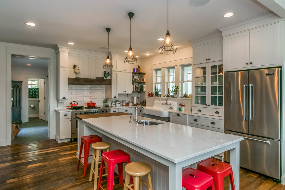 Photo of a country l-shaped kitchen in Cedar Rapids with a farmhouse sink, shaker cabinets, white cabinets, white splashback, subway tile splashback, stainless steel appliances, dark hardwood floors, with island and brown floor.
