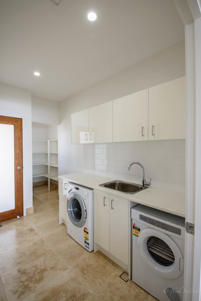 This is an example of a mid-sized contemporary single-wall dedicated laundry room in Brisbane with a single-bowl sink, flat-panel cabinets, white cabinets, white walls, travertine floors and a side-by-side washer and dryer.