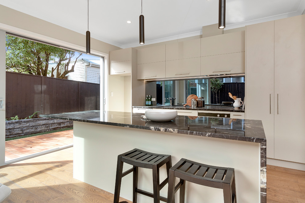 This is an example of a galley open plan kitchen in Auckland with flat-panel cabinets, grey cabinets, marble benchtops, light hardwood floors, with island and black splashback.