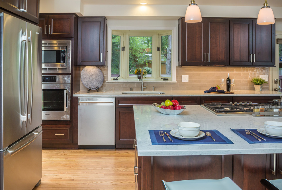 Mid-sized transitional l-shaped light wood floor open concept kitchen photo with an undermount sink, recessed-panel cabinets, dark wood cabinets, quartz countertops, gray backsplash, subway tile backsplash, stainless steel appliances and an island