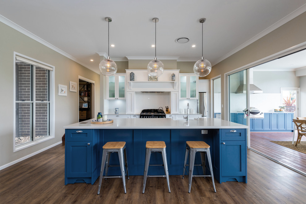 Transitional kitchen in Brisbane with a farmhouse sink, shaker cabinets, blue cabinets, white splashback, black appliances, medium hardwood floors, with island, brown floor and grey benchtop.