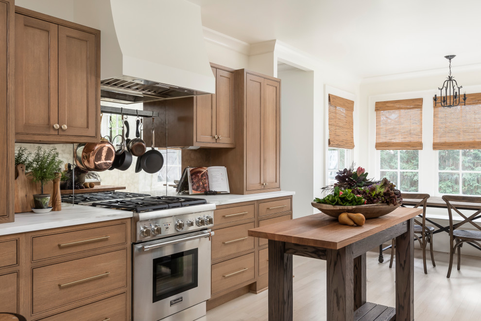Photo of a mid-sized traditional galley separate kitchen in Chicago with a drop-in sink, beaded inset cabinets, medium wood cabinets, marble benchtops, metallic splashback, mirror splashback, stainless steel appliances, light hardwood floors, with island, brown floor and white benchtop.