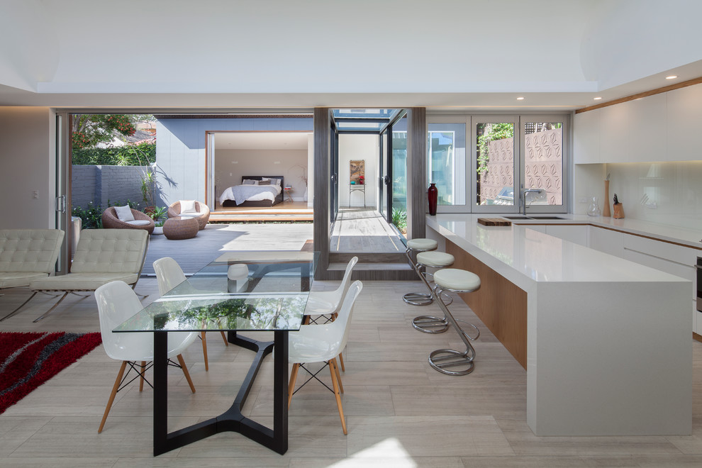 This is an example of a mid-sized modern u-shaped eat-in kitchen in Sydney with a double-bowl sink, white cabinets, quartzite benchtops, white splashback, glass sheet splashback, stainless steel appliances, travertine floors, flat-panel cabinets and a peninsula.