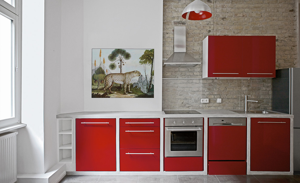 This is an example of a mid-sized eclectic single-wall kitchen in Berlin with a drop-in sink, flat-panel cabinets, red cabinets, concrete benchtops, brown splashback, glass sheet splashback and stainless steel appliances.