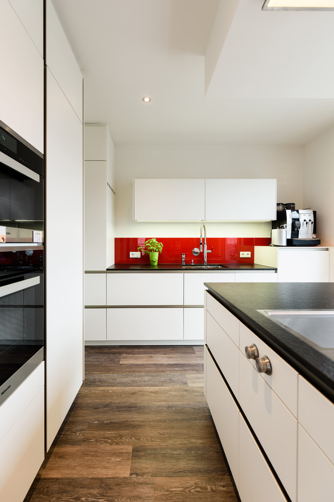 This is an example of an expansive contemporary galley open plan kitchen in Stuttgart with a drop-in sink, flat-panel cabinets, white cabinets, solid surface benchtops, red splashback, glass sheet splashback, black appliances, dark hardwood floors, with island and brown floor.