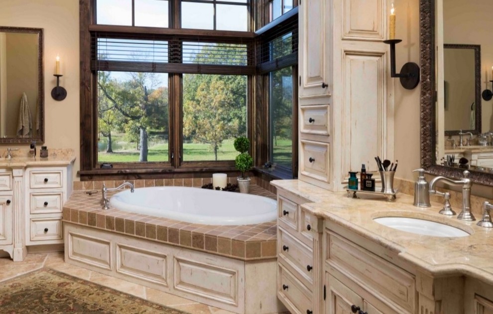 This is an example of a large traditional master bathroom in Los Angeles with louvered cabinets, a corner tub, beige tile, brown tile, granite benchtops, distressed cabinets, ceramic tile, beige walls, travertine floors and an undermount sink.