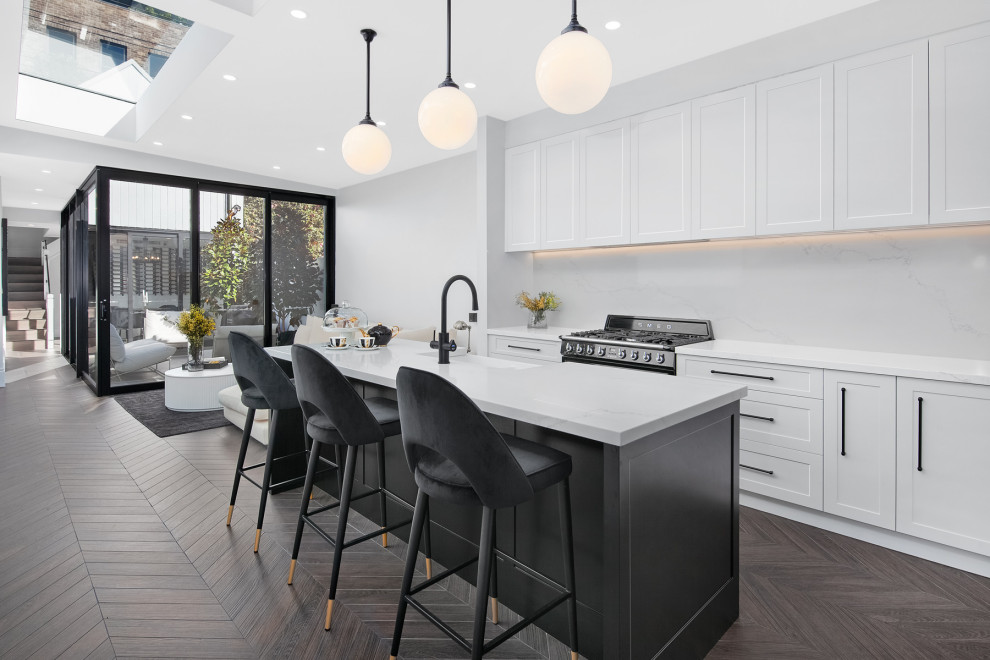 Medium sized single-wall open plan kitchen in Sydney with a belfast sink, shaker cabinets, white cabinets, engineered stone countertops, white splashback, engineered quartz splashback, black appliances, ceramic flooring, an island, brown floors, white worktops and a drop ceiling.