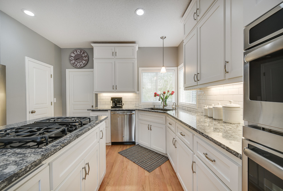 Example of a mid-sized transitional l-shaped light wood floor and brown floor enclosed kitchen design in Portland with a drop-in sink, shaker cabinets, white cabinets, quartz countertops, white backsplash, subway tile backsplash, an island and stainless steel appliances