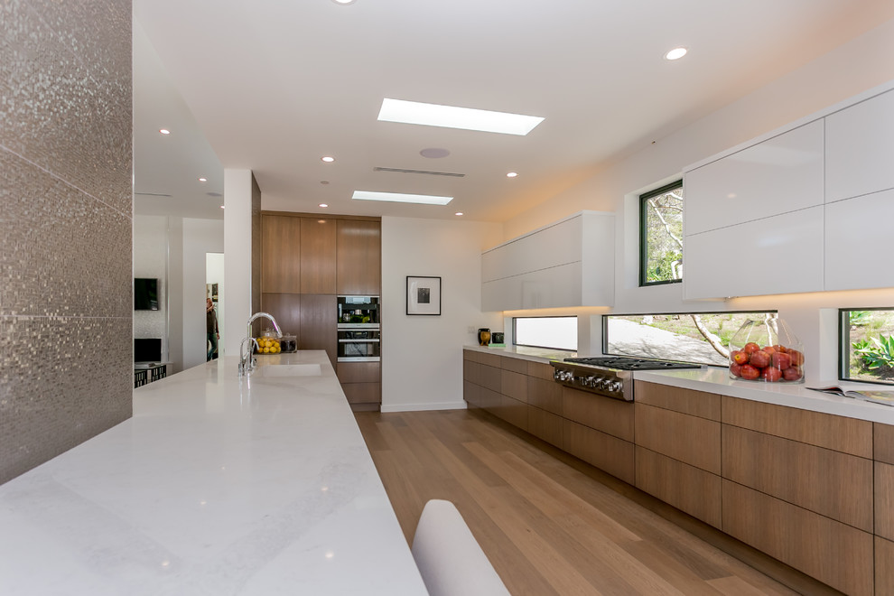 This is an example of an expansive contemporary galley eat-in kitchen in Los Angeles with an undermount sink, flat-panel cabinets, medium wood cabinets, solid surface benchtops, metallic splashback, porcelain splashback, stainless steel appliances, medium hardwood floors and with island.