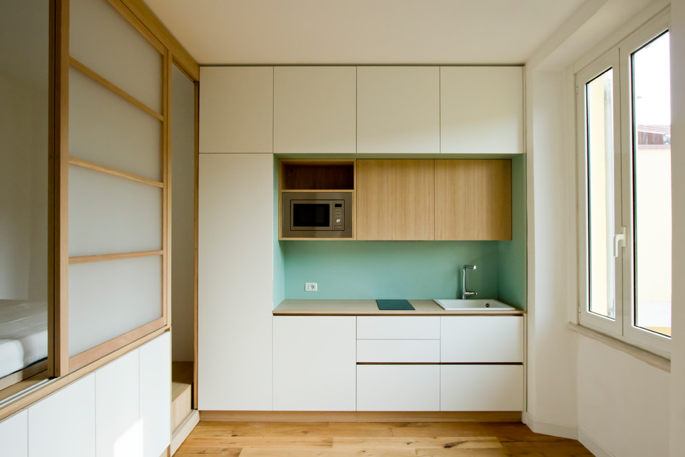 Photo of a small scandinavian single-wall open plan kitchen in Milan with a drop-in sink, flat-panel cabinets, white cabinets, wood benchtops, green splashback, timber splashback, stainless steel appliances, light hardwood floors, brown floor and brown benchtop.