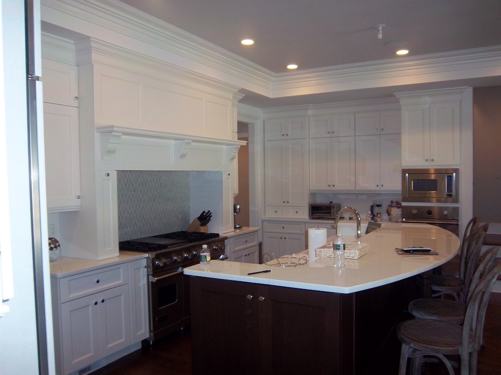 Large transitional l-shaped dark wood floor and brown floor eat-in kitchen photo with a farmhouse sink, shaker cabinets, white cabinets, solid surface countertops, white backsplash, stainless steel appliances and an island
