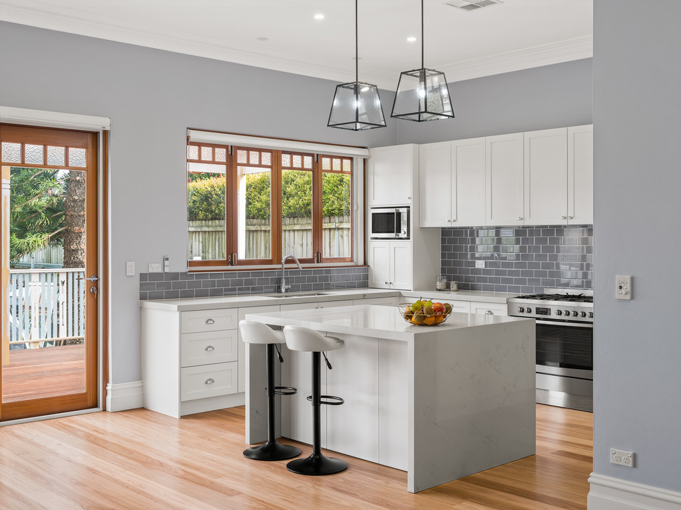 Mid-sized traditional u-shaped open plan kitchen in Sydney with an undermount sink, shaker cabinets, white cabinets, quartz benchtops, grey splashback, ceramic splashback, stainless steel appliances, light hardwood floors, with island, brown floor and grey benchtop.