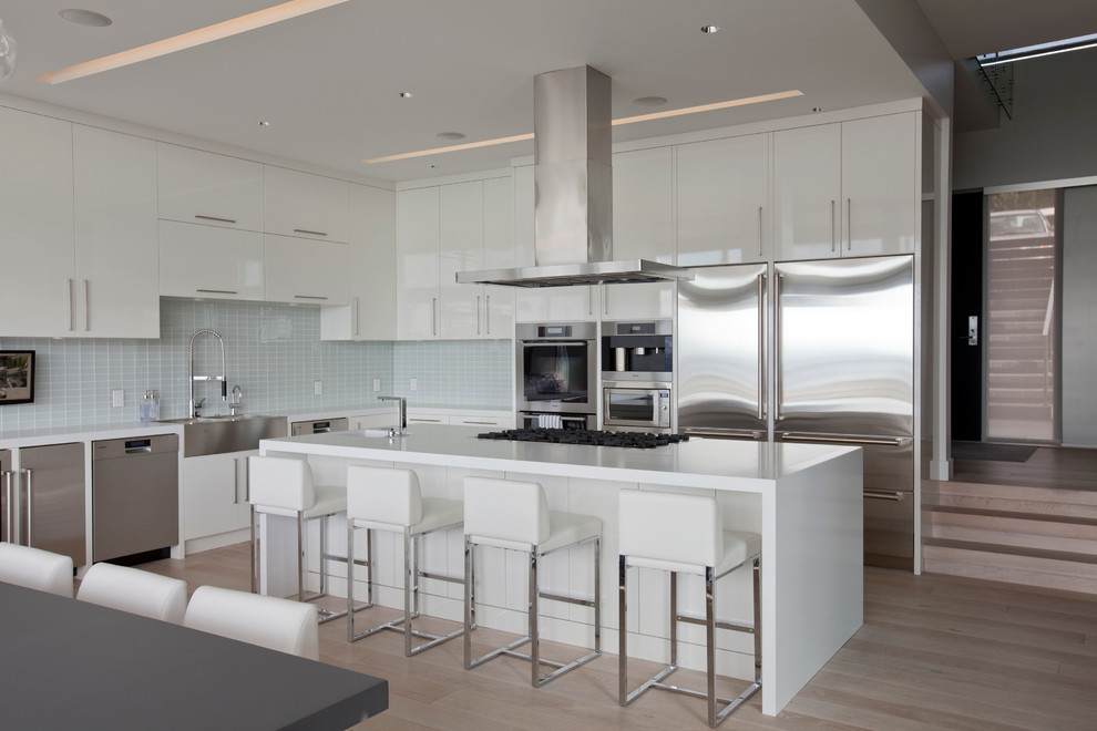 This is an example of a contemporary l-shaped eat-in kitchen in Vancouver with a farmhouse sink, stainless steel appliances, flat-panel cabinets, white cabinets, white splashback, glass tile splashback and quartz benchtops.