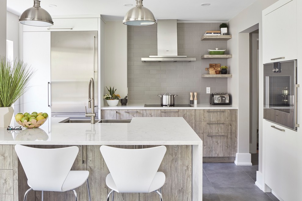 This is an example of a large beach style kitchen in Toronto with a double-bowl sink, flat-panel cabinets, light wood cabinets, marble benchtops, grey splashback, stainless steel appliances, a peninsula, grey floor, white benchtop and subway tile splashback.