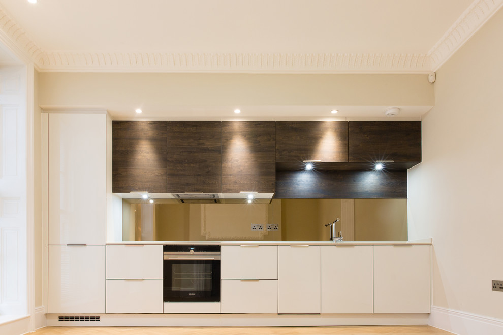 This is an example of a small contemporary single-wall open plan kitchen in London with a drop-in sink, flat-panel cabinets, solid surface benchtops, mirror splashback, black appliances, light hardwood floors, no island and white benchtop.