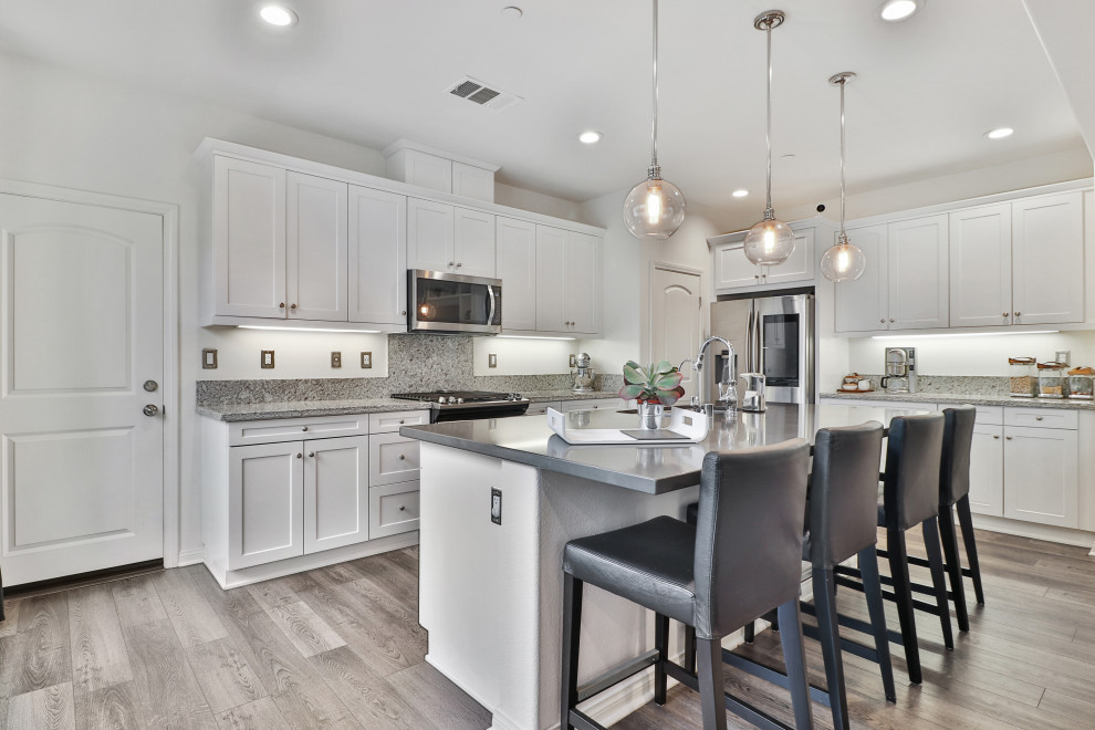 Inspiration for a mid-sized mediterranean l-shaped medium tone wood floor and gray floor open concept kitchen remodel in Los Angeles with a drop-in sink, recessed-panel cabinets, white cabinets, granite countertops, gray backsplash, stainless steel appliances, an island and gray countertops