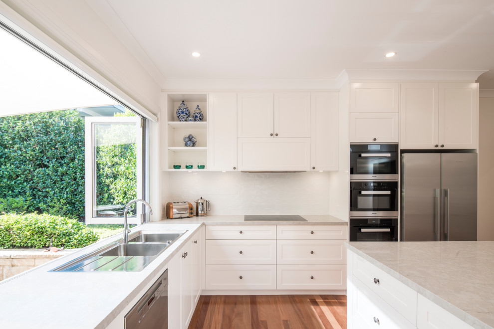 Design ideas for a large transitional l-shaped kitchen in Sydney with a drop-in sink, shaker cabinets, white cabinets, quartz benchtops, white splashback, porcelain splashback, medium hardwood floors, with island, stainless steel appliances, brown floor and grey benchtop.