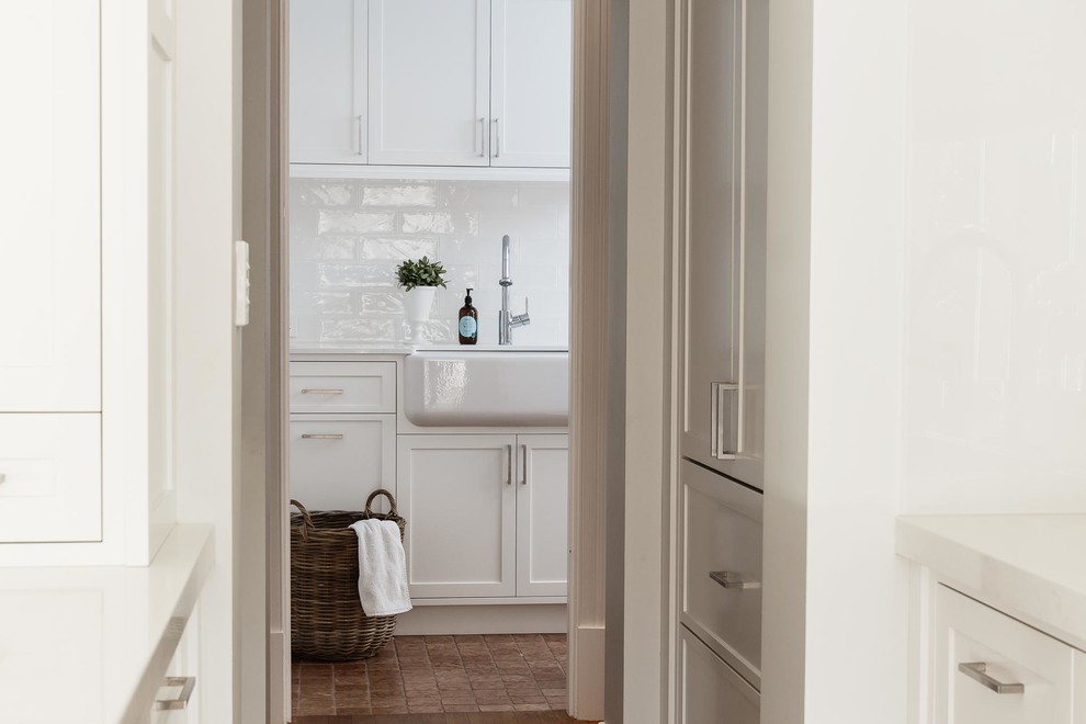 Design ideas for a mid-sized traditional l-shaped laundry room in Perth with a farmhouse sink, shaker cabinets, white cabinets, white walls, porcelain floors, a side-by-side washer and dryer and brown floor.
