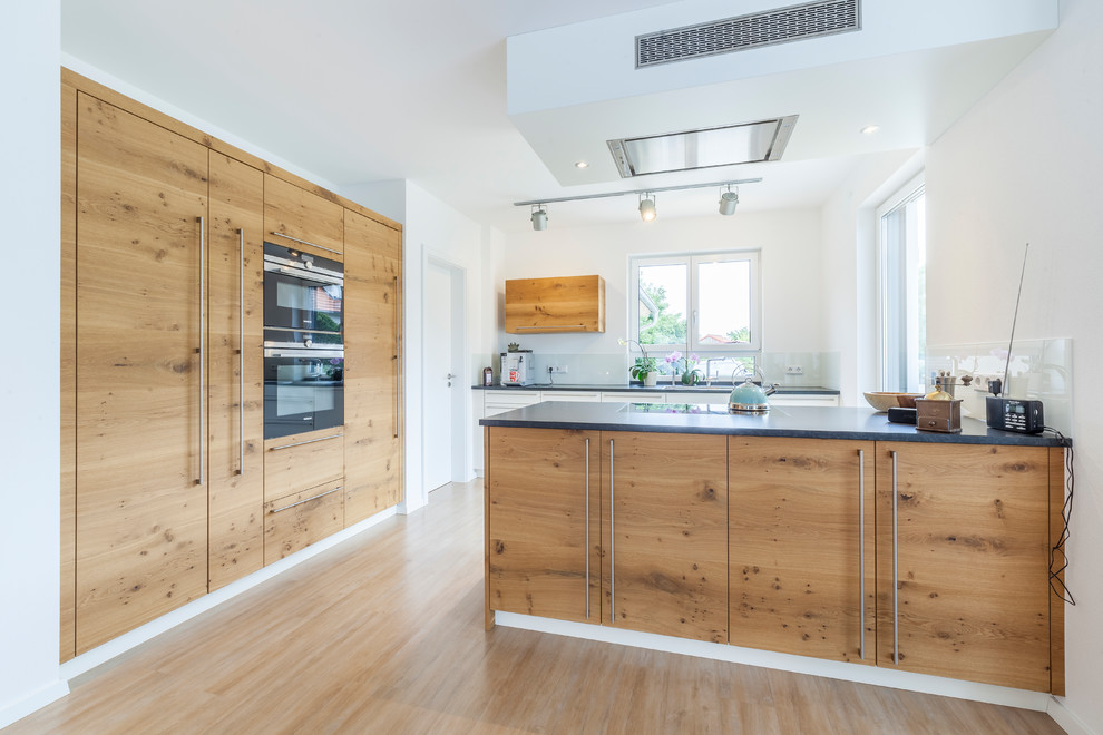 This is an example of a large contemporary l-shaped kitchen in Other with flat-panel cabinets, medium wood cabinets, white splashback, glass sheet splashback, black appliances, light hardwood floors, a peninsula, beige floor and black benchtop.